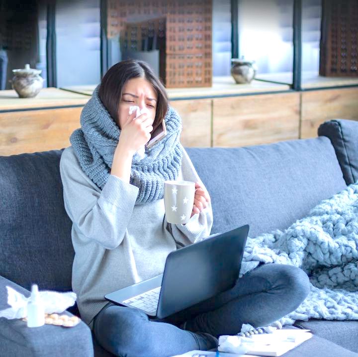 Ill brunette girl is sitting at sofa and working remotely on laptop. Female is blowing out snot, having fever and headache. Young woman is treated at home, took sick leave. Winter cold and flu concept
